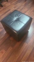 Side angle view of black leather square footstool showing stitched button detail on top and smooth sides.