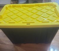 Side and top view of large heavy duty storage container showing yellow lid and black base with ribbed sides on wooden floor indoors.