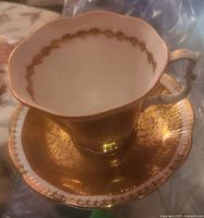 Top and side view of the Royal Albert Buckingham Series teacup on the matching saucer showing gold decorative pattern and scalloped edges.