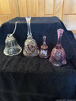 Four vintage glass bells arranged on black cloth backdrop, showing clear, ruby red, cranberry and pink hues with etched and painted designs.