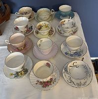 Twelve assorted fine bone china cups and saucers on white surface, with varied floral patterns and gold trim, showing condition and variety.