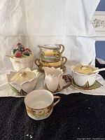 View of all assorted hand painted dishes including bee floral set, stacked cream and sugar dish, cup, and strawberry jam dish with spoon.