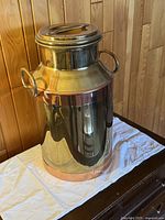 Full view of the brass milk can showing the brass body with copper band near base and copper accent band around handles. Positioned on white cloth on wooden surface against wood panel wall.
