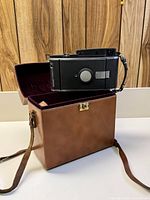 Front side view of Polaroid Model 150 Land Camera placed on the brown leather carrying case with strap visible.