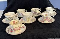 Full set of 8 assorted cups and saucers arranged in two rows on a black cloth background. Each cup has a unique floral design and corresponding saucer.