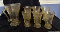 Full set of one yellow Depression glass pitcher and six matching glasses displayed on black cloth backdrop, showing overall condition and style.