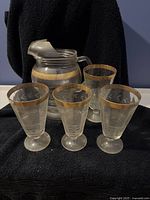 Set of a glass pitcher with etched leaf and stripe design and gold band around the top rim, accompanied by four matching glass drinking glasses with the same etched design and gold band.