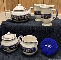 Full view of complete tea set including teapot, two mugs, milk jug, sugar bowl and blue velvet Tetley tin on black cloth