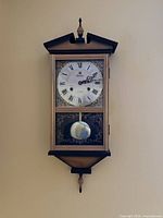 Front view of Luxus wall clock mounted on wall showing decorative wooden case, Roman numeral clock dial, and pendulum chamber.