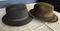 Two fedora hats side-by-side showing a grey Stetson hat with braided grey trim and a brown Stetson hat with braided brown trim on a white surface
