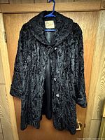 Front view of vintage black Persian lamb fur jacket coat hanging on a hanger showing the wide collar, button closure, and fur texture.