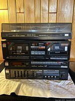 Stacked audio components including turntable on top, double cassette deck, tuner, and amplifier below showing front control panels and dust covers on the turntable.