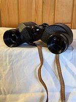 Front view of the textured black Simor Precision binoculars with attached leather strap on soft fabric background.