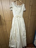 Full view of ivory vintage wedding dress hanging on wooden hanger against wooden wall, showing lace overlay bodice, short sleeves, bow at waist, and skirt detailing.