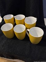 Group of six yellow mugs with white interior and handles arranged on black fabric.