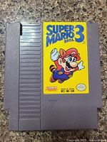 Front view of the Super Mario Bros. 3 NES game cartridge showing colorful label featuring Mario in flying pose on a yellow background.
