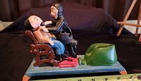 Front and side view of the cast iron dentist coin bank with patient in chair and green coin holder on base, showing size with measuring tape