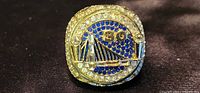 Close-up of front top of large gold-tone championship ring showing Golden State Warriors logo and decorated with blue and clear stones.