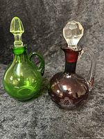 Both decanters shown together on a dark textured surface, displaying their colors and stoppers.