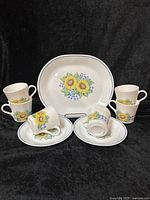 Photo showing six coffee mugs, two luncheon plates, and one serving platter, all featuring sunflower and blue gingham ribbon design on white Corelle dishes.