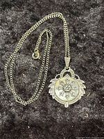 Full view of the silver toned necklace with pendant laid on a dark surface to show the chain and clasp as well as overall length and design.