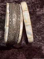 Two vintage bangle bracelets placed vertically on a dark textured background, showing one metal floral engraved bangle and one mother of pearl inlay bangle.