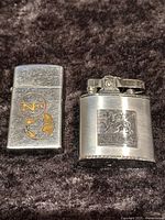 Two vintage metal lighters side by side on dark fabric: a brushed stainless Ronson Triumph lighter with Pegasus design and a Zippo lighter with NF logo and globe.