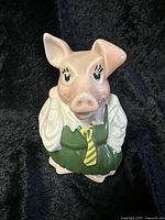 Front and angled view of Wade England pottery piggy bank shaped as a pig character named Girl Annabel. Pig wears a green vest and yellow striped tie over a white shirt, light pink face, approx. 6.5 inches tall.