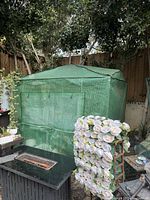 Full view of green mesh-covered greenhouse set outdoors with surrounded plants and garden items.
