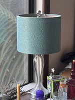 Photo shows a silver metal table lamp with textured blue fabric cylindrical lampshade on a table by window.