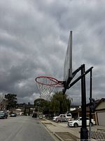 Full view of the basketball hoop from the side showing the hoop, net, pole and backboard under a cloudy sky.