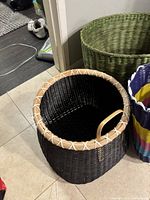 Three woven baskets grouped on a floor in a corner: large green basket, medium black basket with tan rim and handle, and tall multicolored striped basket.