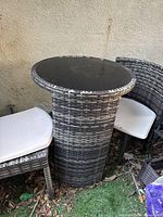 View shows the round nesting table with a dark glass top and wicker base in grey tones alongside two wicker chairs with white cushions.