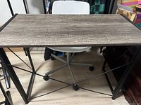 Brown folding desk with wood-textured surface and black metal frame, shown fully opened with white chair behind it.