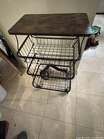 Photo of black metal rolling cart with wooden top and three wire basket shelves on wheels.