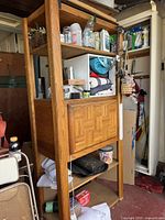 Full view of the oak cabinet showing open shelves with various stored items and the fold down door with a parquet pattern in the front. Visible signs of nail holes and paint spots on the frame.