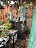 Full view of the brown wicker patio bar with metal topper, surrounded by plants and garden items, showing some wear and outdoor exposure.