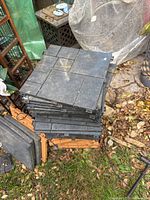 Stack of black plastic patio tiles, showing top textured surfaces resembling pavers, approximately 12x12 inch tiles, used condition with light dirt and wear.
