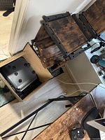 Disassembled brown wood grain shelf panels and grey plastic connectors inside a cardboard box with additional shelf parts leaning against the wall.