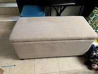 Beige storage ottoman closed, showing fabric texture and rectangular shape with small black feet.