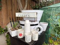 Photo of white garden decor arranged outdoors on a table in front of a wooden fence. Features tall ribbed vase, cylindrical vase with floral pattern, five white pots including one labeled 'Homegrown' with poppy design, square pot with succulents, metal rectangular planter with garden welcome sign, and two large basins.