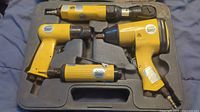 Four yellow air tools laid in molded plastic carrying case showing impact wrench, air ratchet, air drill, and die grinder.