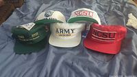Five unworn ballcaps with tags laid out on a bed. Caps include Michigan State, NCSU, Army Lacrosse, and Osagee River themes.
