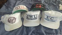 Photo showing five ballcaps arranged in two rows, all caps with tags and featuring collegiate and sports logos.