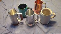 Five assorted beer mugs visible on a fabric surface, showing variety of materials and finishes.