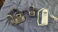 Two Uniden cordless handsets, one with base and answering machine, and one single charger base, alongside white VTech corded phone on a blue surface.