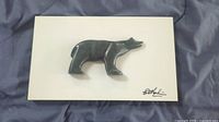 Photo of soapstone bear carving mounted on a flat white backing, showing the entire mounted piece with visible hand-signed mark on bottom right corner of the mounting.