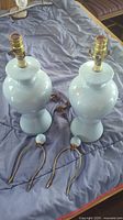 Pair of matching light blue ceramic table lamps with cords and finials shown on a bed