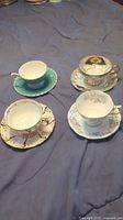 Four assorted porcelain teacups with matching saucers shown together on blue cloth background. Each set has distinct decorative patterns and gold accents.