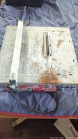 Full view of the tile saw showing metal base, adjustable guide bar, and power cord, with rust spots visible on the surface.